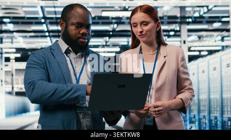 Doing maintenance, laptop and engineering team in server hub working together to finish tasks. Configuring settings, servers and teamworking colleagues in modern high tech data center, camera B Stock Photo