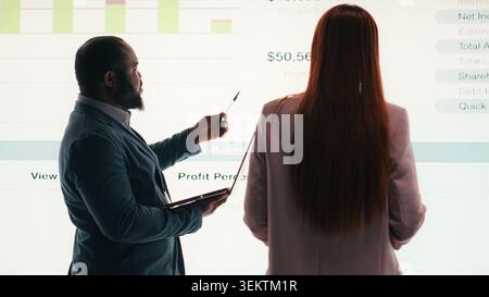 Analyzing economic data graphs, laptop and businesspeople using dashboard, brainstorming. verifying company financial statements, charts projected on digital wall, and teamworking colleagues, camera B Stock Photo