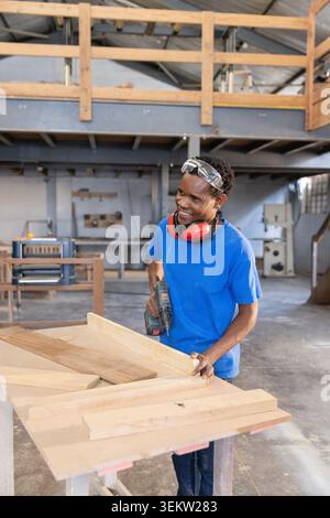 African American male carpenter drilling plank at bench in carpentry shop using cordless drill Stock Photo