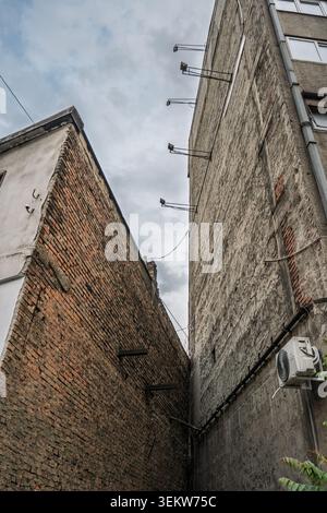 Low-angle view looking up between two tall building facades, showing a narrow shadowy gap, weathered brick walls, concrete surfaces, metal brackets an Stock Photo
