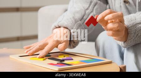 Senior woman solving tangram puzzle at home, elderly hands arranging colorful wooden pieces on board for brain training, memory improvement, cognitive Stock Photo