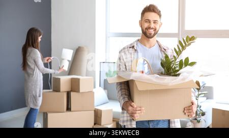 Smiling young man holding moving box with houseplant and home decor while woman unpacks cardboard boxes in bright new apartment, happy couple relocati Stock Photo