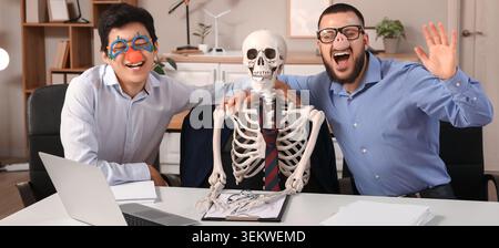Funny office coworkers posing with skeleton colleague at desk, cheerful business team in silly party glasses and pig nose celebrating halloween in mod Stock Photo