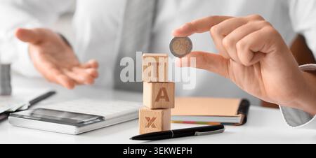 Hand placing euro coin on wooden tax block stack with calculator, paperwork and blurred businessperson in office, concept of taxation, tax payment, fi Stock Photo