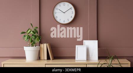 Minimalist home interior with wall clock above wooden cabinet, potted houseplant, books and empty photo frames on warm brown wall Stock Photo