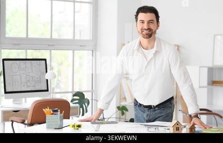 Smiling architect standing at desk with house plans in bright modern office, confident interior designer or engineer portrait in creative workplace wi Stock Photo
