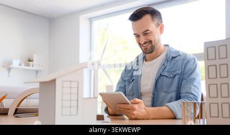 Smiling architect or engineer using tablet at desk with house model and wind turbine in bright modern office, sustainable building design, renewable e Stock Photo