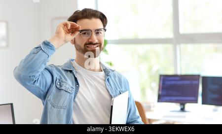 Smiling young man in eyeglasses holding laptop and adjusting glasses in bright modern office, portrait of confident programmer, developer or office wo Stock Photo