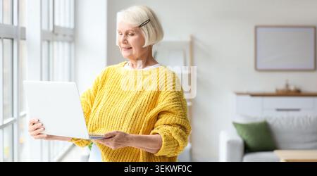 Senior woman using laptop at home near window, smiling mature female in yellow sweater working online, browsing internet, remote communication, digita Stock Photo