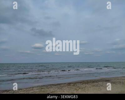 A wide view of gentle ocean waves rolling onto a quiet sandy shore under a vast, overcast gray sky filled with low-hanging clouds during a somber, pea Stock Photo