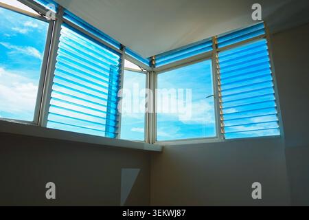 Interior corner view looking up at bright blue glass louvered windows against a clear blue sky, casting geometric natural light and shadows on white walls Stock Photo