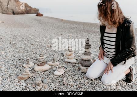 Stones beach meditation woman kneeling on a pebbly shore by the sea building rock cairns for mindfulness and tranquility Stock Photo
