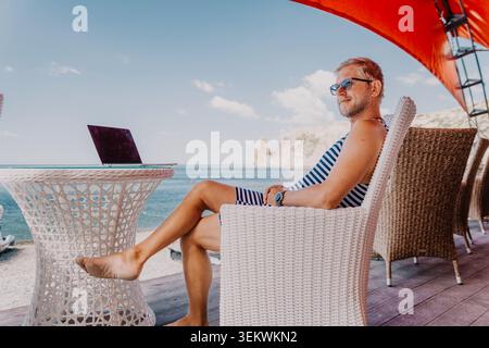Man laptop beach mature traveler sitting in a wicker chair at a coastal cafe working on his computer with an ocean view Stock Photo