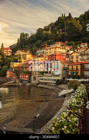 Varenna, Italy - 11.10.2022: Vibrant waterfront houses perched on a steep green slope above a peaceful pebble shore with blooming flowers Stock Photo