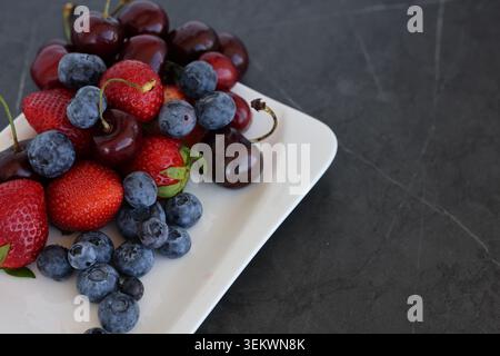 Fresh Mixed Berries and Cherries on White Plate Over Dark Background. A colorful assortment of fresh strawberries, blueberries, and cherries Stock Photo