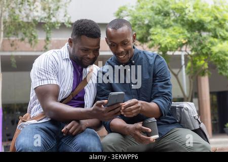 Two African men sitting on low concrete bench, leaning in, sharing smartphone and coffee Stock Photo