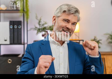 Middle-aged businessman at home office dances on chair wearing earphones singing rock song after working on laptop. Freelancer guy at table writes code enjoying music energy boosting high productivity Stock Photo