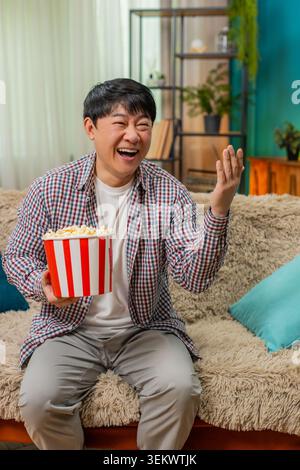 Asian man laughs loudly watching comedy movie, holding stomach and eating popcorn, sitting on home sofa. Chinese guy enjoys humorous entertainment, relaxed joyful reaction fills room with carefree fun Stock Photo