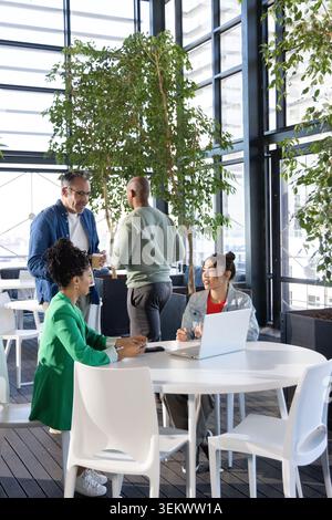 Diverse coworkers wearing business attire meeting around round table in office atrium with laptop Stock Photo
