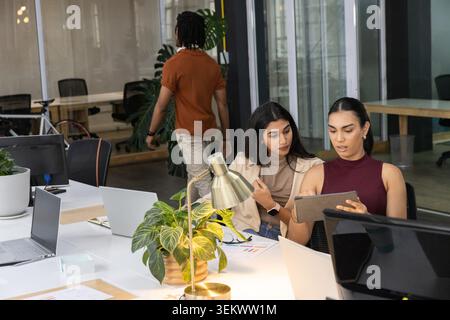 Diverse coworkers two adult women in blazers leaning in examining tablet near brass lamp in office Stock Photo