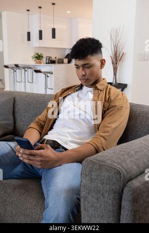Adult male using smartphone while sitting on gray sofa in modern living-room with open-plan kitchen Stock Photo