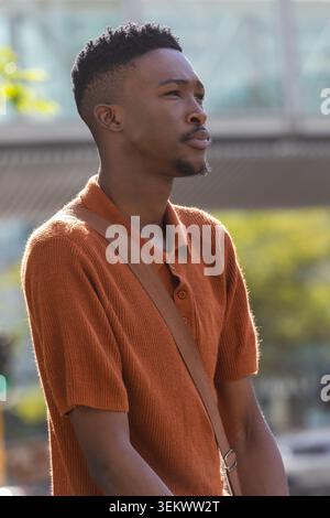 African man standing gazing right in sunny plaza wearing orange polo, crossbody strap, lip piercing Stock Photo
