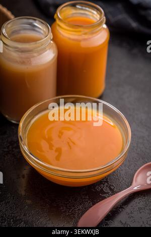 Creamy pumpkin soup in a glass bowl surrounded by jars of puree and a spoon, warm orange tones suggesting homemade autumn comfort, healthy vegetarian Stock Photo