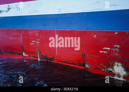 Scratched and scuffed red and blue painted hull of a large cargo vessel at the water surface, close-up industrial view. Stock Photo