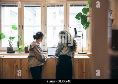 Rear view of female business expert standing beside coworker using laptop at table in new tech company Stock Photo