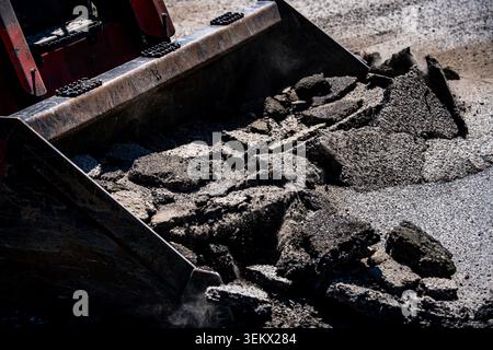 Excavator bucket moves broken asphalt. Heavy excavator supports road repair. Excavator works on construction site. Damaged pavement needs excavator Stock Photo