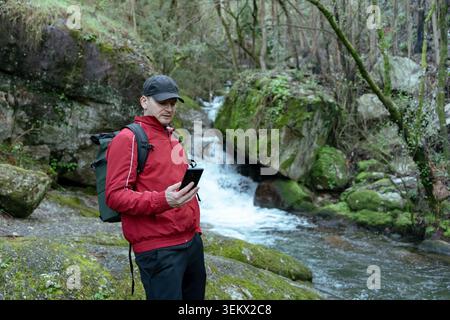 Man using smartphone for navigation during a hike in a mossy forest with a waterfall Stock Photo