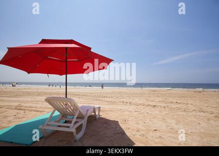 A red parasol and white sunbed on a sandy beach under a clear blue sky, capturing a relaxing midsummer vacation with the calm ocean background. Stock Photo