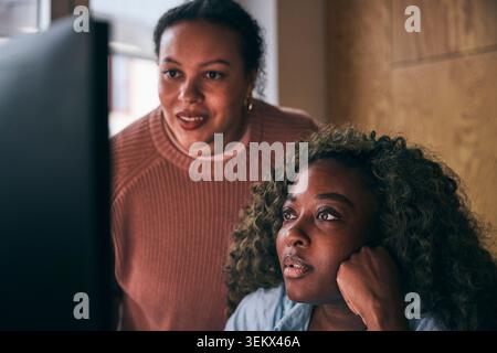 Focused curly haired businesswoman analyzing data on computer with female coworker in tech office Stock Photo