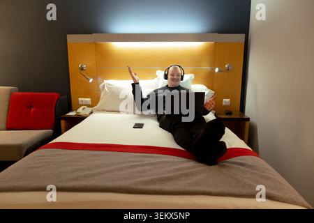 Young adult man relaxing on hotel bed, working remotely with laptop and headphones Stock Photo