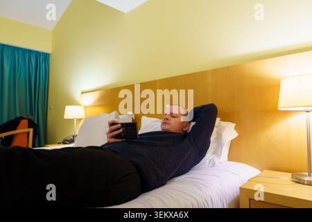 Man lying on bed in a hotel room, watching content on phone Stock Photo