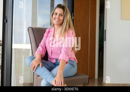 Portrait of a happy woman sitting comfortably on a chair in a bright modern home. Lifestyle image representing wellbeing, confidence, relaxation, posi Stock Photo