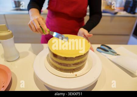 Hands applying creamy icing to a freshly baked cake using a spatula in a kitchen Stock Photo