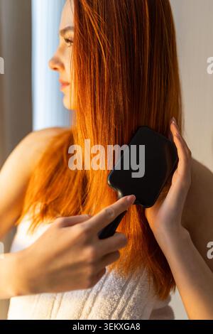 Young woman brushing long red hair after shower in morning sunlight Stock Photo