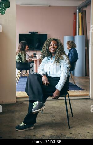 Portrait of confident curly haired female entrepreneur sitting on chair in tech hub Stock Photo