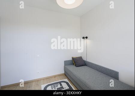 minimalist room with a gray sofa, white walls, a round ceiling light, and a play mat Stock Photo