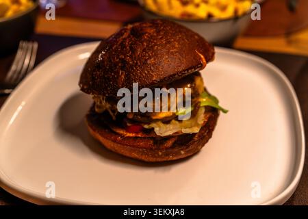 Close view of gourmet cheeseburger with dark bun served on a white plate in a restaurant with warm indoor lighting. Stock Photo