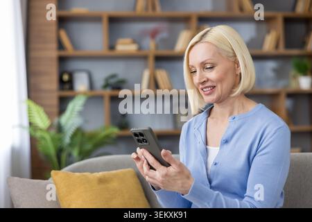 Cheerful blonde woman sitting on a sofa and happily scrolling through content on a modern smartphone, connecting with digital technology and online lifestyle Stock Photo