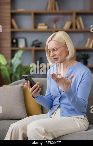 Frustrated middle-aged woman sitting on a sofa at home, looking at her smartphone with a confused and annoyed expression, feeling overwhelmed by technology issues Stock Photo