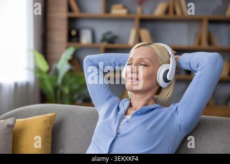 Senior woman enjoying peaceful moment listening to music with eyes closed and white headphones on, relaxing with hands behind head on a cozy sofa in her living room Stock Photo