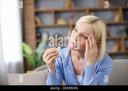 Mature blonde woman suffering from severe migraine and stress, touching her head with a hand, holding a white tissue, looking sad and tired, sitting indoors on a couch Stock Photo