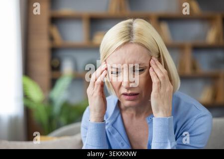 Senior woman suffering from a severe headache or migraine, pressing temples with hands, feeling intense pain, stress, and exhaustion while sitting indoors Stock Photo