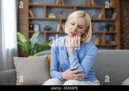 Mature blonde woman feeling sharp tooth pain, sitting on a sofa at home and pressing her hand against her jaw due to discomfort and oral health problems Stock Photo