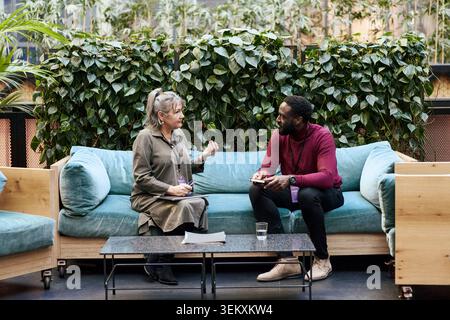 Senior female business expert discussing with male associate while sitting on sofa in convention center lobby Stock Photo