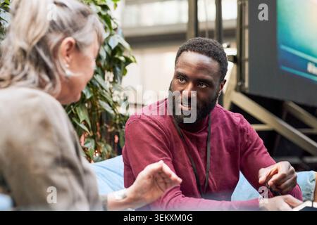 Male business expert discussing with senior female associate while sitting on sofa in convention center lobby Stock Photo