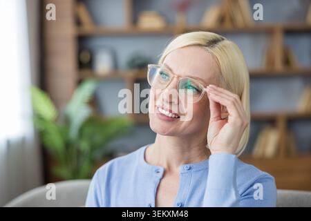 Blonde woman adjusting her eyeglasses while smiling, looking up and to the side, expressing inspiration, wisdom, and deep thought in a home office setting Stock Photo